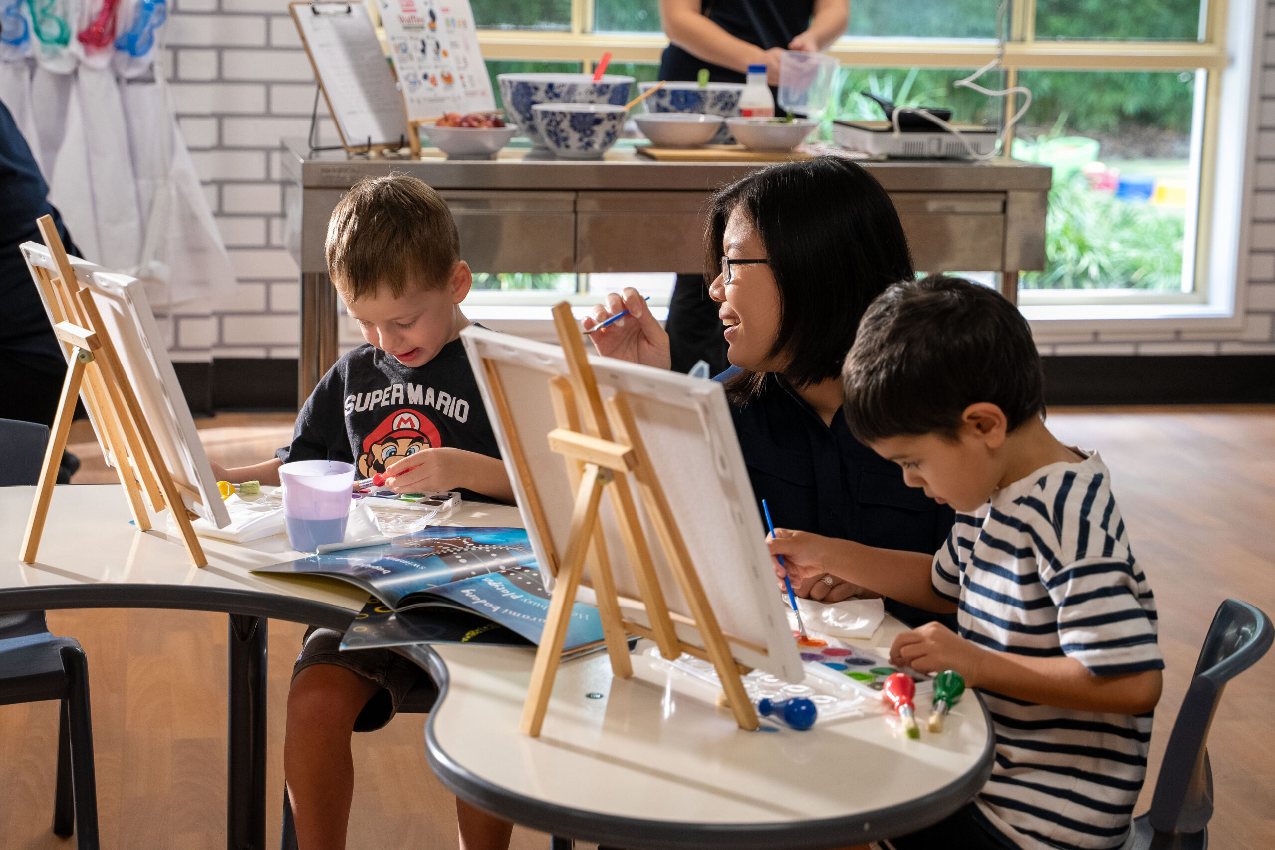 An Imagine Childcare educator supports two children painting at tabletop easels