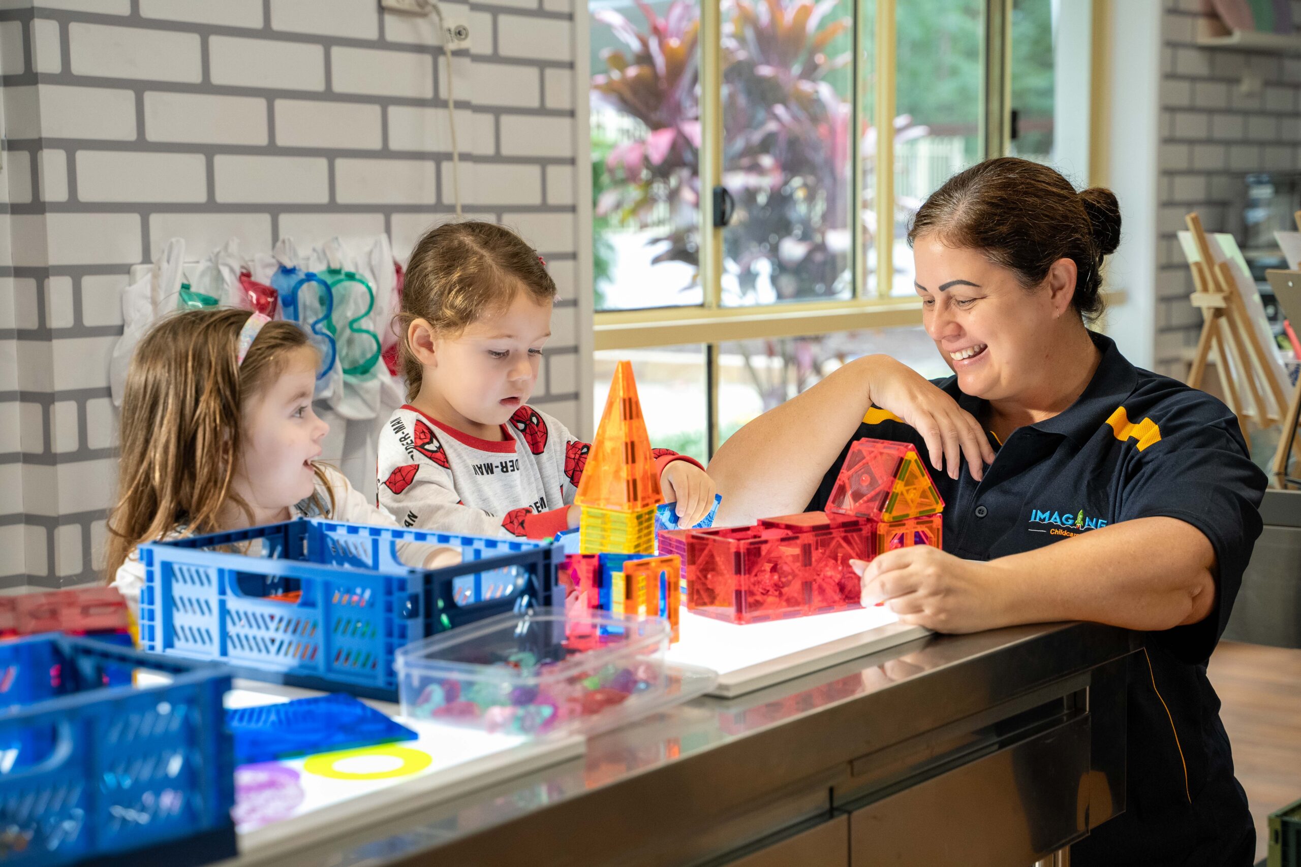 Children and an Imagine Childcare educator build colourful magnetic tile structures on a light table